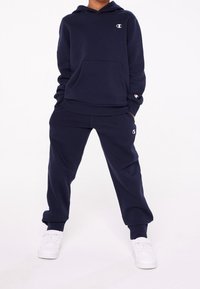 Felpa blu navy con tasca frontale e logo, abbinata a pantaloni coordinati. Entrambi realizzati in tessuto morbido; presentano polsini e vita elastici.