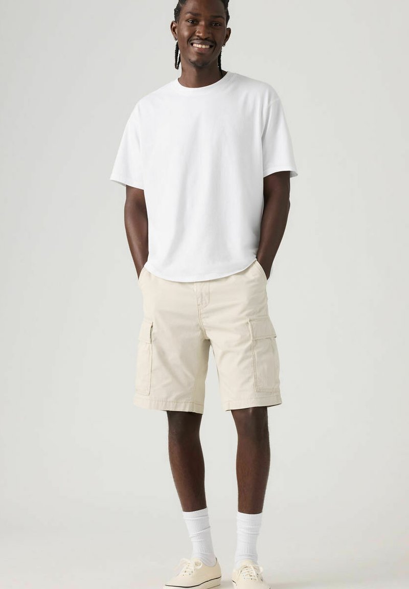 T-shirt en coton blanc, coupe décontractée, associé à un short cargo beige clair avec poches latérales, longueur genou, porté avec des chaussettes blanches et des chaussures claires.