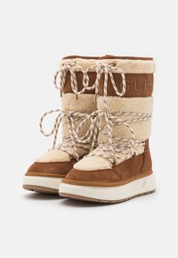 Botas de ante marrón y lana de oveja crema con suelas gruesas beige, que presentan cordones trenzados y herrajes en tono dorado, logo grabado en la parte superior.
