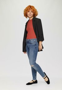 Blazer noir, haut couleur rouille, jeans déchirés bleus et chaussures plates noires. Cheveux bouclés orange. Design simple et moderne sans motifs visibles.