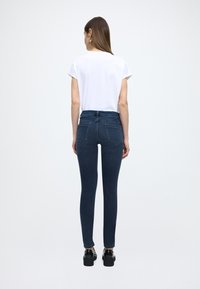 Tumši zili, pieguļoši džinsi ar augstu vidukli, kombinēti ar vienkāršu baltu t-kreklu. Melni masīvi apavi. Modelis redzams no muguras.