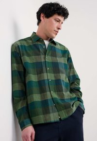 Green plaid shirt with a button-down collar, featuring varying shades of green and blue, made of textured fabric, worn over a white shirt.