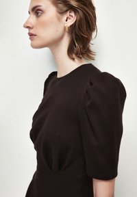 Tmavě hnědress má nabírané rukávy, kulatý výstřih a boční kapsu. Modelka má na sobě zlaté kruhové náušnice. Viditelná hladká textilní struktura.