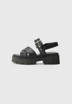 Schwarze Plateausandalen mit einer klobigen Sohle, breiten Kreuzriemen und einem verstellbaren Knöchelriemen mit einer Schnalle und metallischen Akzenten.