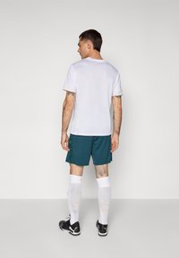 Witte kortemouwenshirt, teal korte broek met groen patroon, witte kniesokken, zwarte sportschoenen, gezien vanuit de rug.