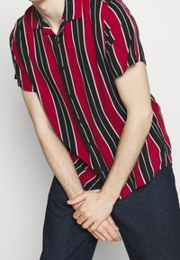 Osoba w czerwono-czarnej koszuli w pionowe paski z krótkim rękawem, trzymająca ręce złożone przed ciemnoniebieskimi spodniami na białym tle.