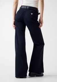 Granatowe dzwony jeansowe z talią ozdobioną wzorem, dwoma tylnymi kieszeniami i małą naszywką z logo nad prawą kieszenią.