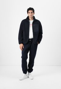 Jachetă din polar albastru închis cu un buzunar frontal, combinată cu pantaloni jogger albaștri închis și adidași albi. Textură netedă și închidere cu fermoar, cu guler înalt.