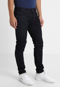 Tmavě modré džíny se slim fit střih, s kontrastním šitím a designem se pěti kapsami. Nosené s bílými teniskami.