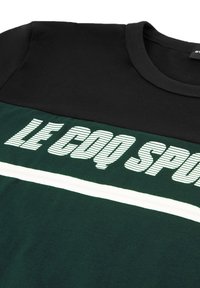 Camiseta verde y negra con rayas horizontales. Texto blanco "LE COQ SPORTIF" en una fuente atrevida y rayada en la parte frontal. Material de algodón.