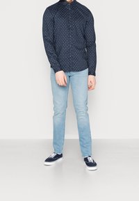 Man som bär en marinblå långärmad skjorta med små vita mönster, ljusblå jeans och marinblå sneakers med vita snören.