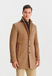 Brązowy płaszcz z wełny z wysokim kołnierzem, zapięciem na pojedynczy guzik i kieszeniami po bokach, nałożony na pikowaną ciemną kurtkę. Gładka tekstura.