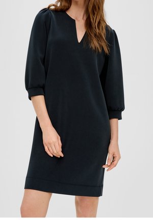 Kobieta ubrana w czarną sukienkę za kolano z bufkami na rękawach 3/4 i dekoltem w kształcie litery V, stojąca na tle jednolitego jasnego tła.