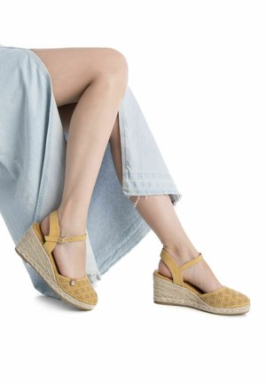 Sandali con zeppa perforata gialla e cinturino alla caviglia, suola intermedia rivestita in fibra di juta, punta rotonda e superficie testurizzata. Indossati con una gonna di jeans azzurra chiara.