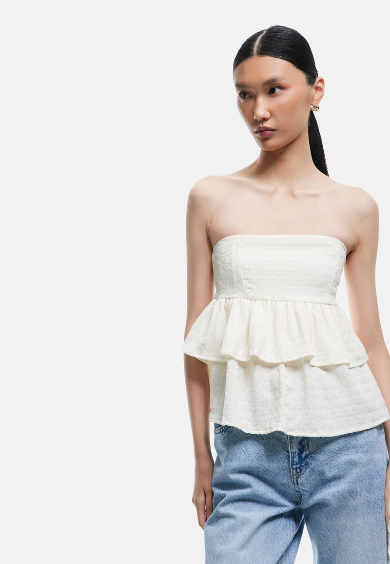 Hvid peplum top uden stropper med struktureret stof, der har en flagrende kant og lagdelt flæse-design, parret med lyseblå denim jeans.