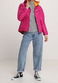 Różowa pikowana kurtka z ociepliną o wydzielonym wzorze, z pomarańczowym wnętrzem, noszona na szarej bluzie z zamkiem i jasnoniebieskich dżinsach.