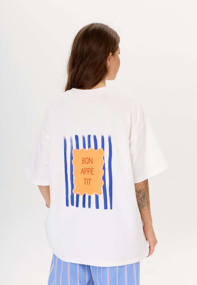Weißes übergroßes T-Shirt mit einem orangefarbenen Grafikdesign und blauen gestreiften Akzenten sowie dem Text "BON APPÉTIT" in fetter Schrift auf der Rückseite.