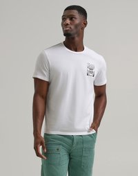 Uomo in t-shirt bianca con grafica di una scatola di Crayola e pantaloni verdi con grandi tasche, in piedi contro uno sfondo grigio uniforme.
