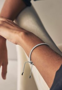 Bracelet ajustable en argent avec un design texturé et deux extrémités à glissière sur un poignet de teint clair, sur un fond neutre.