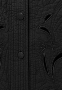 Abito nero strutturato con ricami floreali intricati, linee di cucitura visibili e bottoni a pressione neri lungo la parte anteriore.