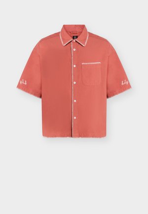 Lyhythihainen korallinvärinen paita, jossa valkoiset siksakommaukset kauluksessa, rintataskussa, hihoissa ja napilliskaitaleessa, varustettu rintataskulla ja kirjotuilla hiha-yksityiskohdilla.