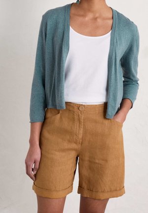 Personne portant un haut blanc, un cardigan bleu canard à manches trois-quarts, et un short marron taille haute avec ourlet roulé et poches.
