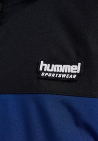 Patch Hummel Sportswear sur une surface en tissu noir et bleu. Le patch présente un texte noir sur fond blanc, embossé avec un contour.