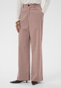 Hlače širom nogavica od mekog ružičastog materijala s glatkom teksturom, s prednjim zatvaranjem na gumb i bočnim džepovima. Uparene s cipelama s oštrim nosom.