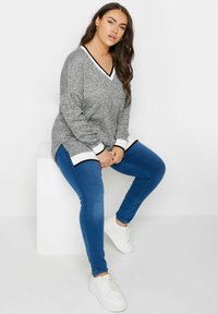 Pullover grigio con texture, colletto a V bianco e nero, abbinato a jeans skinny blu e sneakers bianche. Posizione seduta su un cubo bianco.