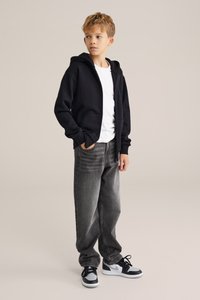 Fekete cipzáras kapucnis pulóver egy fehér póló felett, szürke farmernadrággal és fekete-fehér sneaker cipővel párosítva. Az anyag puhanak és laza stílusúnak tűnik.