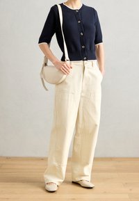 Navy knitted cardigan with short sleeves and five buttons, paired with cream wide-legged trousers and beige flat shoes. Rounded handbag.