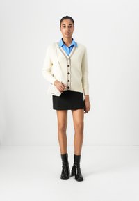 Cream cardigan with textured knit, brown buttons, and contrasting collar; paired with a light blue shirt, black skirt, and black ankle boots.