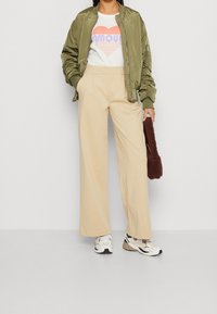 Olivgrön bomberjacka, vit t-shirt med en flerfärgad hjärt-grafik, beige vida byxor och vita sneakers. Brun texturerad handväska.