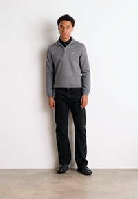 Grauer Strickpullover mit einem Reißverschlusskragen, kombiniert mit schwarzen Jeans. Das Model steht vor einem weißen Hintergrund und trägt schwarze Schuhe.