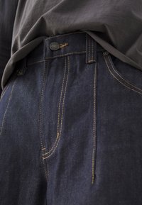Dark denim jeans with yellow stitching, featuring a button closure and front pockets, paired with a gray fabric shirt.