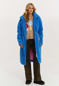 Žena vo forme modrého teddy kabáta cez sveter s farebnými hubami, v nohaviciach s leopardím vzorom a čiernych platformových topánkach, stojaca proti bielej pozadí.