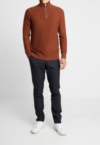 Brun stickad tröja med halvdragkedja, matchad med marinblå randiga byxor och vita sneakers. Texturerad design på tröjan.