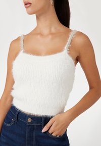 White fuzzy crop top with thin, embellished straps. Soft texture and ribbed pattern. Paired with dark denim, showcasing a high-waisted design.