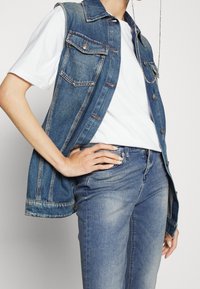 Chaleco de mezclilla con cuello, dos bolsillos en el pecho y cierre de botón, sobre una camiseta blanca y combinado con jeans de color azul claro.