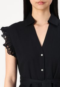 Melna kleita ar apkakli un pogu aizdari. Ar frillēm apšūptām piedurknēm un mežģīņu apdari, kā arī auduma jostu viduklī. Mīksts, teksturēts materiāls.