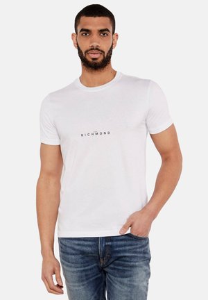 Camiseta de algodón blanca con cuello redondo, mangas cortas y el texto impreso "RICHMOND" en negro. Combinada con jeans de mezclilla azul.