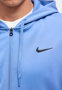 Gros plan d'une personne portant un sweat à capuche zippé bleu clair avec un logo Nike swoosh noir sur la poitrine et un t-shirt blanc en dessous.