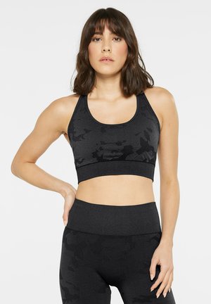 Frau mit schulterlangem dunklem Haar, die einen schwarzen Sport-BH mit Camouflage-Muster und passende hoch taillierte Leggings vor einem einfarbigen Hintergrund trägt.