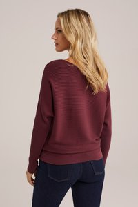 Bordo spalvota megztinis su ilgomis rankovėmis, apvaliu iškirpimu ir prigludusia apačia, derinamas su tamsiai mėlynomis džinsomis. Matoma tekstūruota paviršiaus detalė.