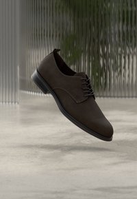 Καφέ οξφορντ παπούτσι από σουέντ με λεία υφή, στρογγυλεμένη μύτη και σχέδιο με κορδόνια. Διαθέτει μαύρη καουτσούκ σόλα και διακριτικό τακούνι.