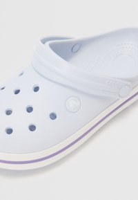 Zoccoli azzurri chiari in plastica con fori di ventilazione, suola bianca con una striscia accentata viola e un logo circolare sul lato.