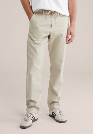 Mand iført beige snore-løse chinos med sidelommer og hvide sneakers med sorte striber, stående foran en neutral baggrund.