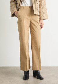 Tan wide-leg trousers made from a smooth fabric, featuring a tailored fit, front pockets, and a classic button closure. Paired with black heeled boots.