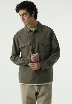 Man draagt een groene overhemdjas met knopen en twee borstzakken, beige broek, kijkt opzij met handen voor zich geklemd.