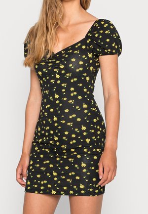 Femme portant une robe noire cintrée avec des manches bouffantes et un petit imprimé floral jaune, debout devant un fond clair uni.